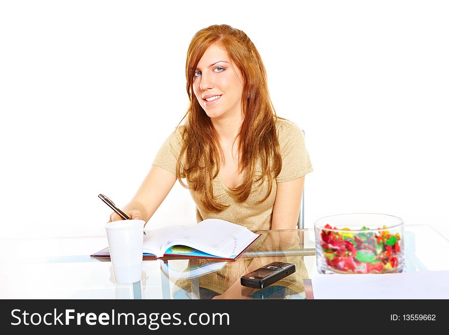 Smiling student girl learning at desk with notebook with pen in hand, mobile phone and some sweets. Smiling student girl learning at desk with notebook with pen in hand, mobile phone and some sweets