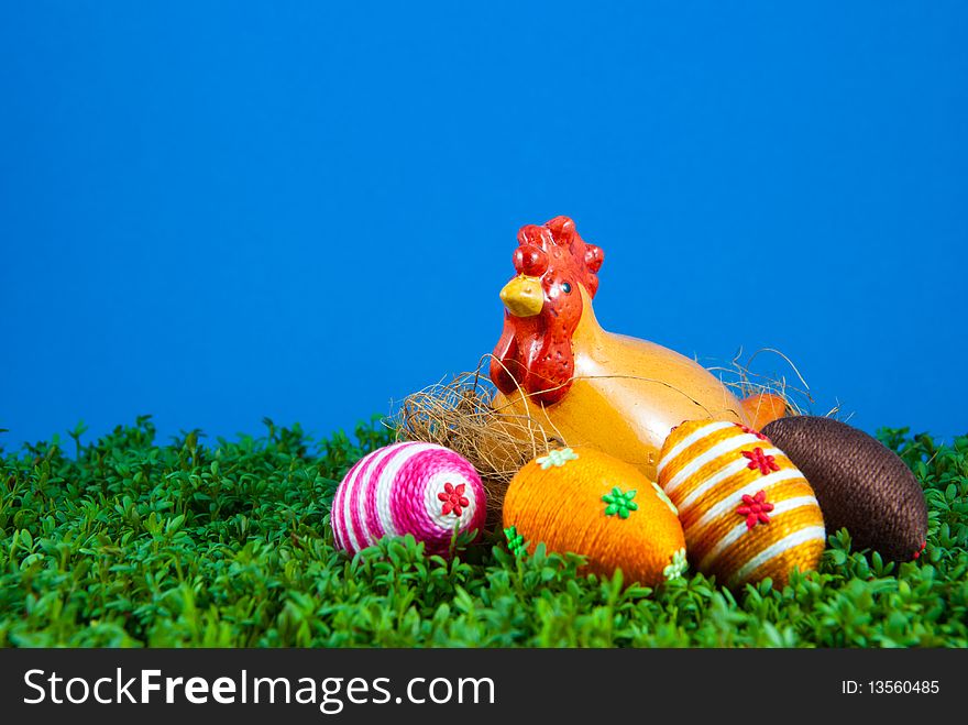 Handmade easter egg and ceramic hen placed on cress. Small eggs are covered by cotton string in different colours. Additional decorative elements are glued to egg. Blue background. Ceramic hen is used as Easter decoration. Handmade easter egg and ceramic hen placed on cress. Small eggs are covered by cotton string in different colours. Additional decorative elements are glued to egg. Blue background. Ceramic hen is used as Easter decoration