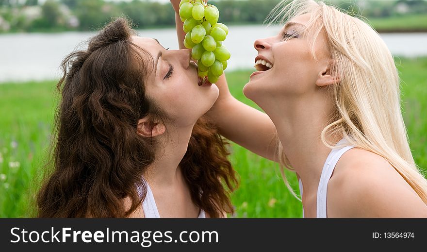 Portrait of two beautiful girls blonde and brunette in white suits, feed each other grapes. Portrait of two beautiful girls blonde and brunette in white suits, feed each other grapes