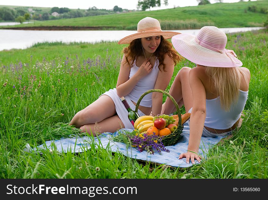 Two attractive girls in hats blonde and a brunette sitting on the grass near the basket with fruit. Two attractive girls in hats blonde and a brunette sitting on the grass near the basket with fruit