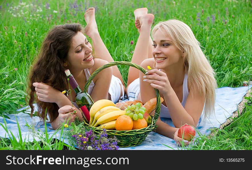 Two attractive girls lie on the rug on the grass beside baskets of fruit and talking. Two attractive girls lie on the rug on the grass beside baskets of fruit and talking