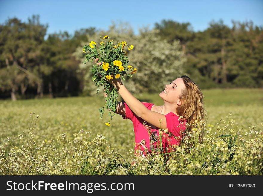 Beautiful young blonde woman standing in blooming meadow in spring, bunch of yellow flowers in hand, smiling, blue sky and trees in background; shallow depth of field. Beautiful young blonde woman standing in blooming meadow in spring, bunch of yellow flowers in hand, smiling, blue sky and trees in background; shallow depth of field