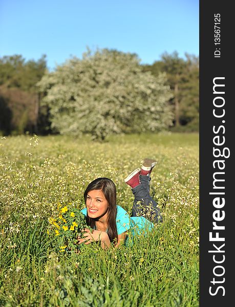 Beautiful young blonde woman lying in blooming meadow in spring, bunch of yellow flowers in hand, smiling, blue sky and trees in background; shallow depth of field. Beautiful young blonde woman lying in blooming meadow in spring, bunch of yellow flowers in hand, smiling, blue sky and trees in background; shallow depth of field