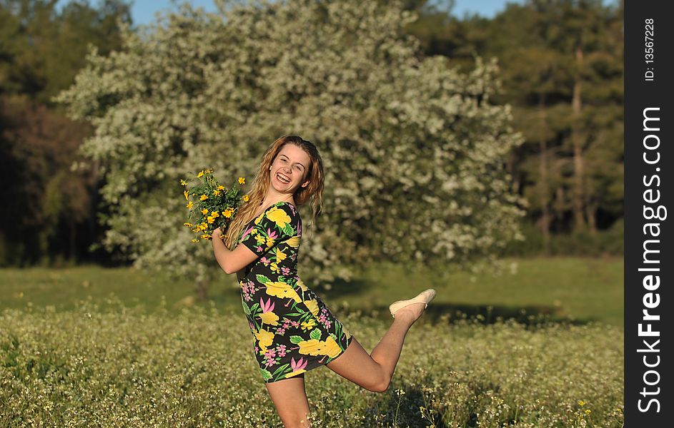 Beautiful young blonde woman jumping in blooming meadow in spring, bunch of yellow flowers in hand; shallow depth of field, trees in background. Beautiful young blonde woman jumping in blooming meadow in spring, bunch of yellow flowers in hand; shallow depth of field, trees in background