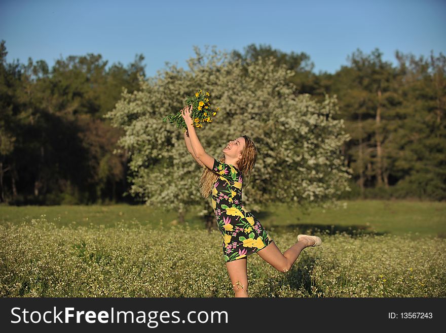 Beautiful young blonde woman jumping in blooming meadow in spring, bunch of yellow flowers in hand; shallow depth of field, trees in background. Beautiful young blonde woman jumping in blooming meadow in spring, bunch of yellow flowers in hand; shallow depth of field, trees in background