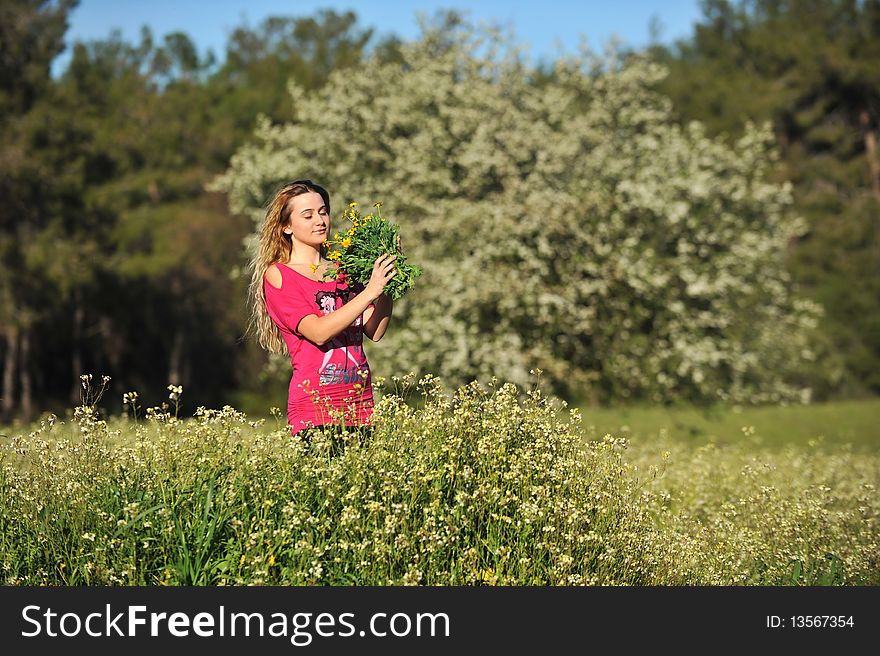 Beautiful young blonde woman standing in blooming meadow in spring, bunch of yellow flowers in hand, smelling eyes closed, blue sky and trees in background; shallow depth of field. Beautiful young blonde woman standing in blooming meadow in spring, bunch of yellow flowers in hand, smelling eyes closed, blue sky and trees in background; shallow depth of field