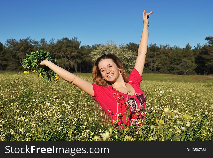 Beautiful young blonde woman standing in blooming meadow in spring, bunch of yellow flowers in hand, cheerful, smiling, blue sky and trees in background; shallow depth of field. Beautiful young blonde woman standing in blooming meadow in spring, bunch of yellow flowers in hand, cheerful, smiling, blue sky and trees in background; shallow depth of field