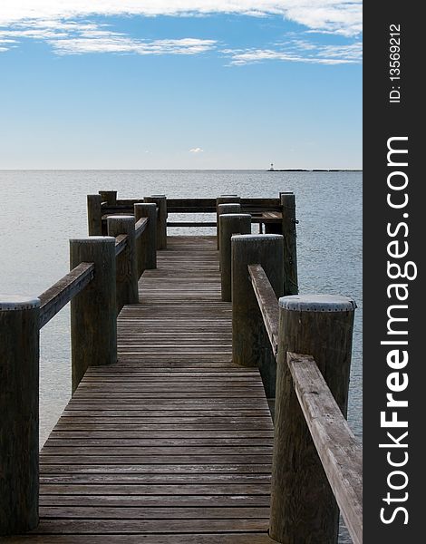 A narrow wooden pier juts out into the ocean on a sunny day with blue sky and sparse white clouds.