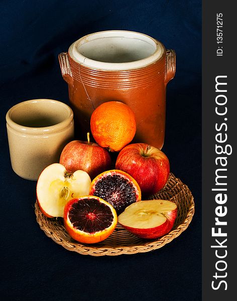 A still life image of two terracotta jars, one cracked, with a wicker tray of fruit, apples and oranges, placed in front. All layed on a navy blue fabric background. A still life image of two terracotta jars, one cracked, with a wicker tray of fruit, apples and oranges, placed in front. All layed on a navy blue fabric background