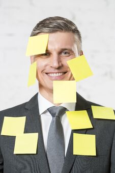Portrait Of Smiling Mature Businessman With Blank Yellow Adhesive Notes On Face And Suit At Office Stock Photo