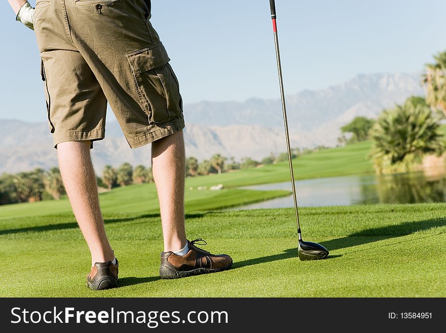 Golfer standing on golf course holding club, (low section)