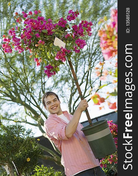 Young man holding small flowering tree in pot in garden centre, smiling