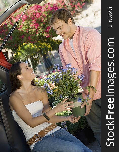 Young couple with potted flower in garden centre, woman sitting in open car trunk
