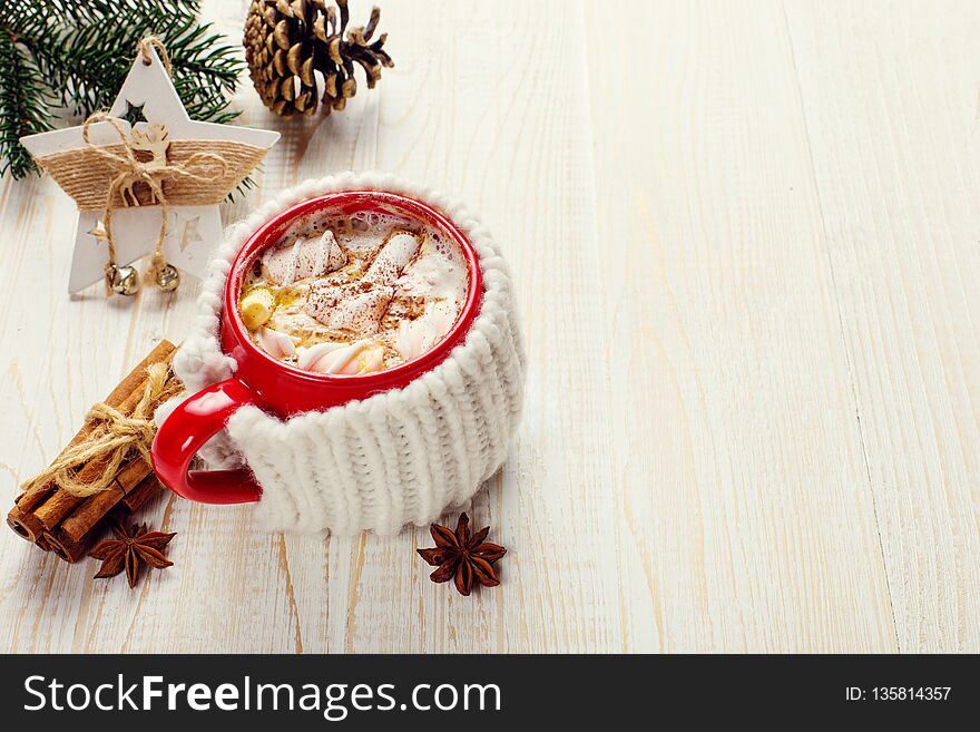 Winter coffee drink, cocoa with whipped cream and marshmallows in a red ceramic cup. It stands on a white wooden table, next to a checkered scarf, a Christmas tree, a pine cone, a star decoration. Place for text, copy space.