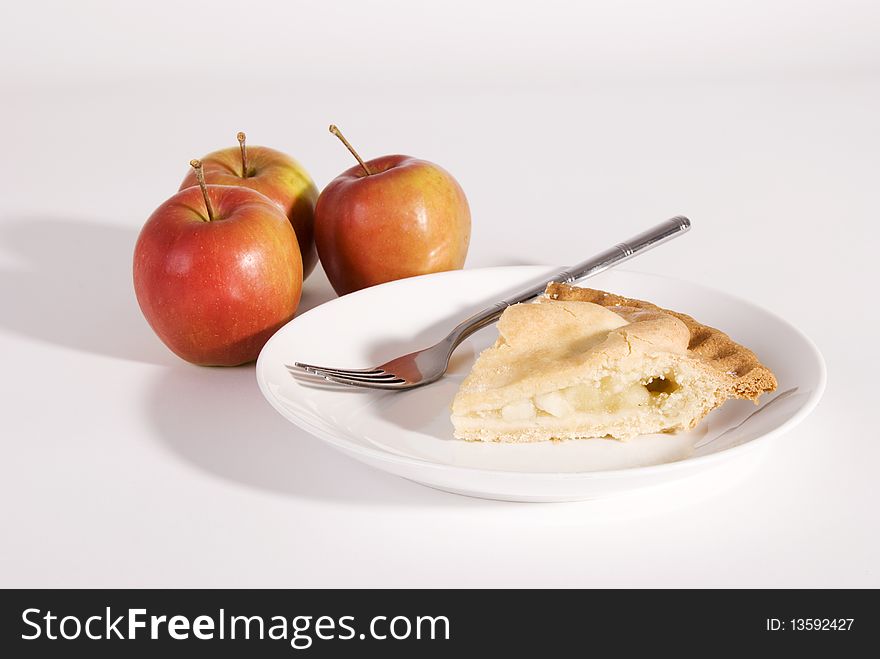 A horizontal image of a slice of apple pie on a white plate with three red apples in the background