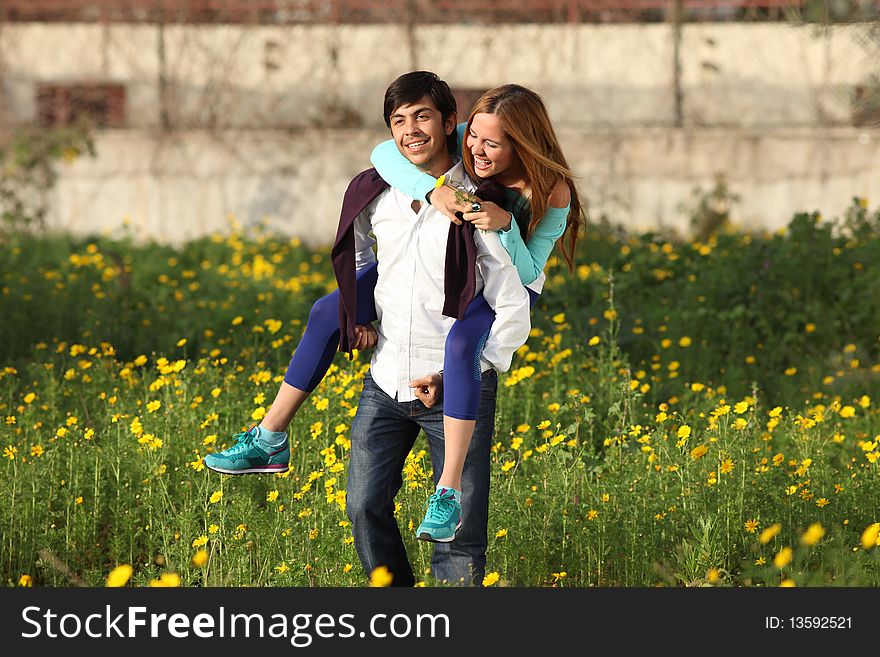 Beautiful young blonde woman with her boyfriend piggy-backing in blooming meadow in spring, yellow flowers in hand, smiling; shallow depth of field. Beautiful young blonde woman with her boyfriend piggy-backing in blooming meadow in spring, yellow flowers in hand, smiling; shallow depth of field
