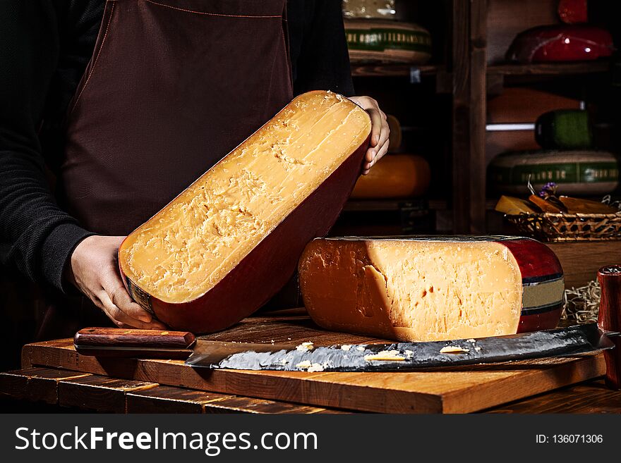 Young Worker Holding Half Of Wheel Gauda Cheese In Shop. Wooden Background, Knife For Cheese. Close Up View. Copy Space For Text