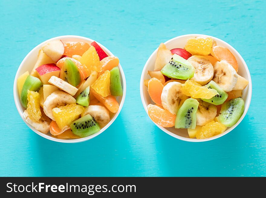 Two white bowls of healthy fresh fruit salad on turquoise background.