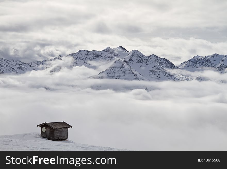 Wooden cabin above the clouds overlooking snowy mountain peaks with a cloudy sky