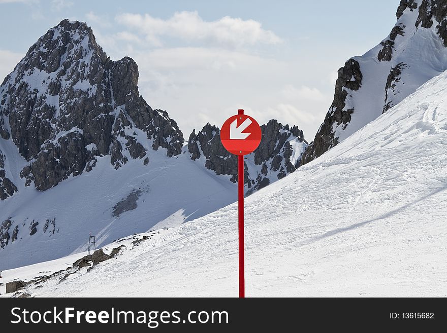 Red sign with arrow pointing down showing direction of ski run with snowy mountain peaks behind. Red sign with arrow pointing down showing direction of ski run with snowy mountain peaks behind