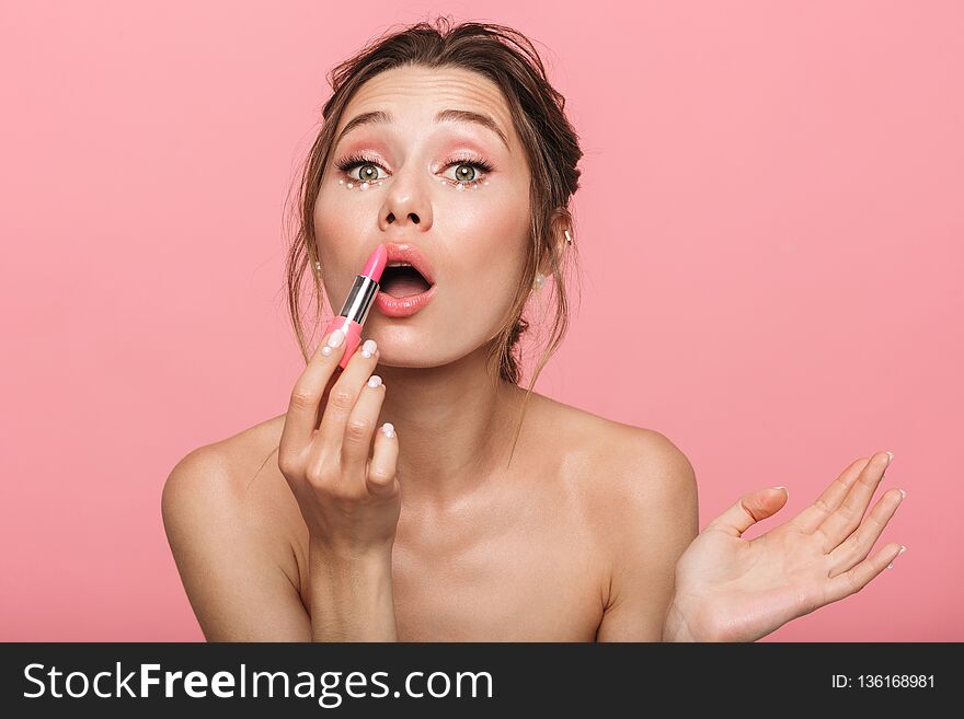Pretty young woman posing isolated over pink wall background holding lipstick doing makeup