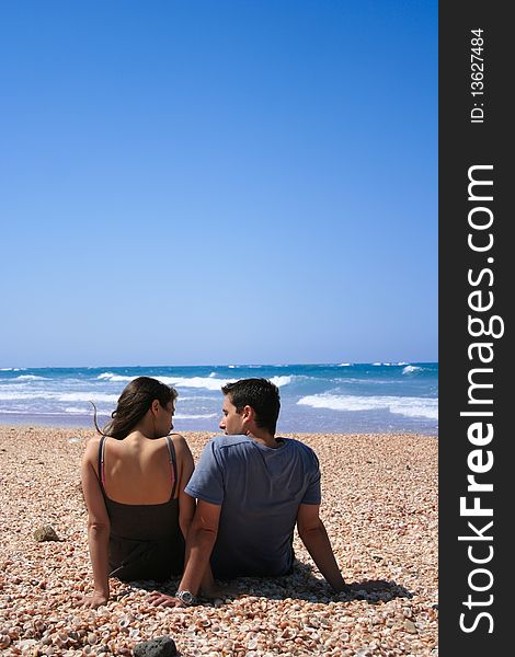 Couple sitting on the beach looking at each other clear blue sky