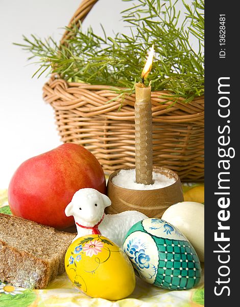 Easter eggs lay on a napkin near bread of a lamb and a burning candle against a basket with green branches. Easter eggs lay on a napkin near bread of a lamb and a burning candle against a basket with green branches