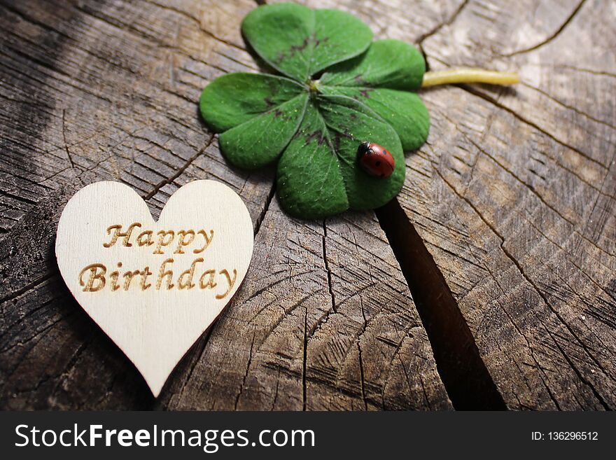 The picture shows a wooden heart with happy birthday greetings and lucky clover with a ladybird.