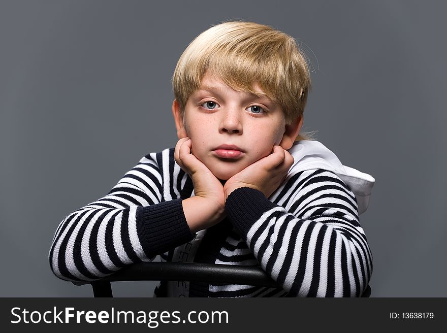 Portrait of a pensive boy in a striped sweater, sitting on a chair on a gray background. Portrait of a pensive boy in a striped sweater, sitting on a chair on a gray background