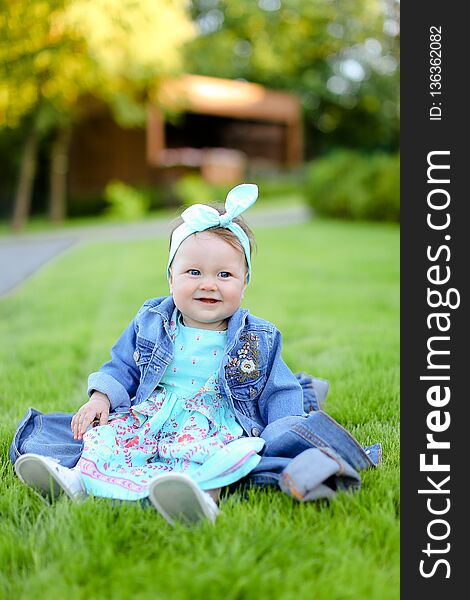 Little happy female child sitting on green grass and wearing jeans jacket.