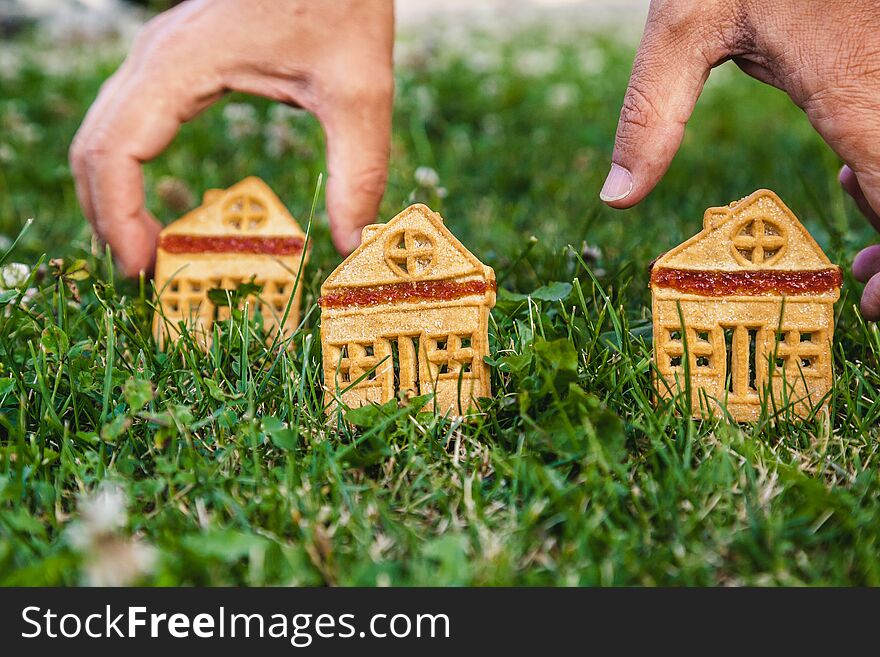 Fabulous sweet house. Three cookies in the form of a house stand on a green meadow close-up. The hand of man puts one house. Fabulous sweet house. Three cookies in the form of a house stand on a green meadow close-up. The hand of man puts one house