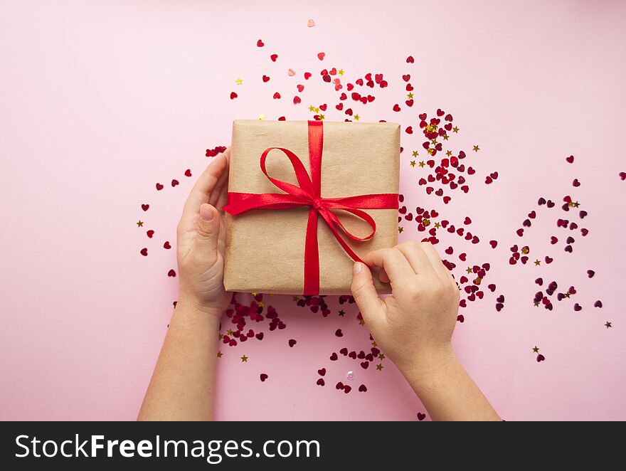 Child is holding gift box wrapped in brown colored craft paper and tied with red bow on pink background with red heart shape confetti. Child is holding gift box wrapped in brown colored craft paper and tied with red bow on pink background with red heart shape confetti.