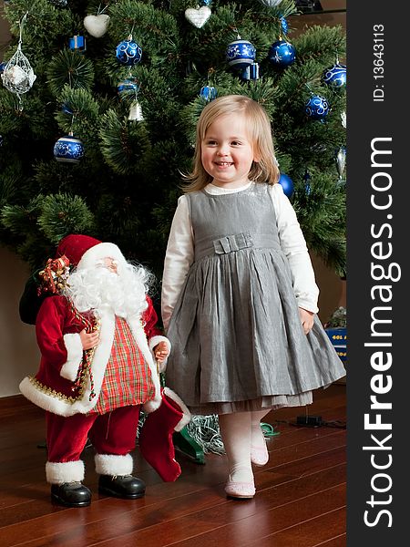 Cute little girl in gray dress with toy Santa Claus under the Christmas tree. Cute little girl in gray dress with toy Santa Claus under the Christmas tree