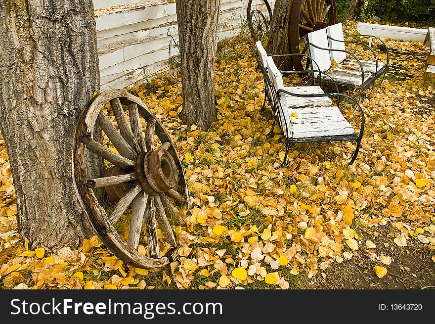 Old weathered wooden wagon wheel leaning against tree trunk with benches in BG and yellow leaves on groound. Old weathered wooden wagon wheel leaning against tree trunk with benches in BG and yellow leaves on groound.