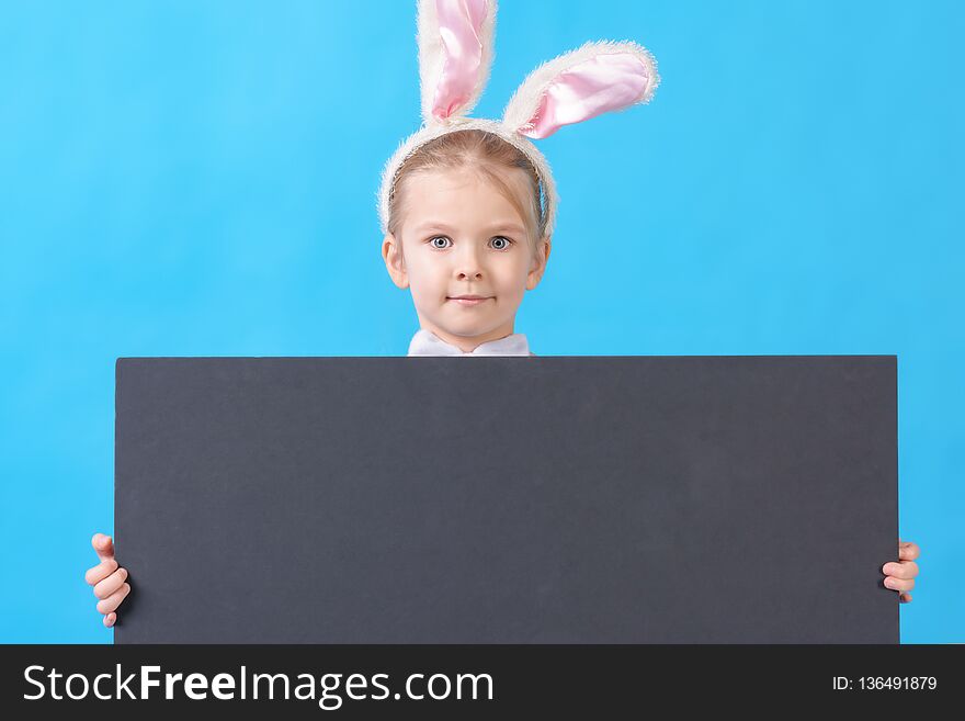 A child in a white rabbit costume on a blue background. A cute little girl with ears of a hare holds in her hands an empty sheet of black cardboard. Advertising photo with space for text.