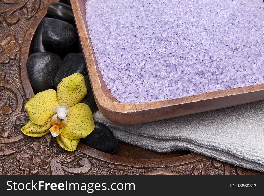Spa scene with bath salts, massage stones, towel and an orchid on in a carved wood setting.