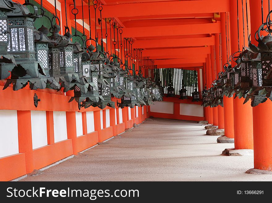 Two rows of traditional Japanese Lanterns line a wall at a temple in Nara, Japan. The scene is very Japanese in style, with red of the temple contrasting with the greenish hue of the lanterns.