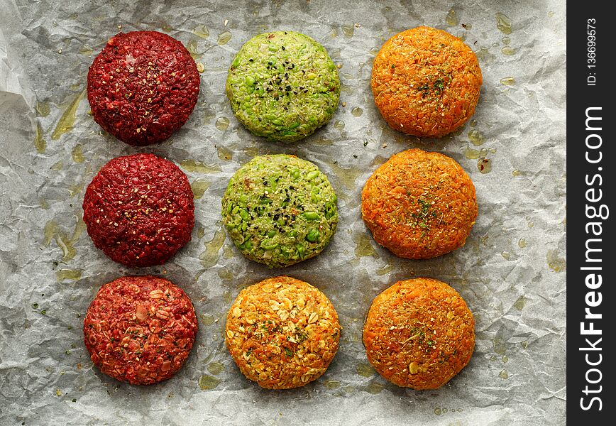 Raw vegan burgers made of beetroot, green peas, carrots, groats and herbs on white parchment prepared for baking, top view.