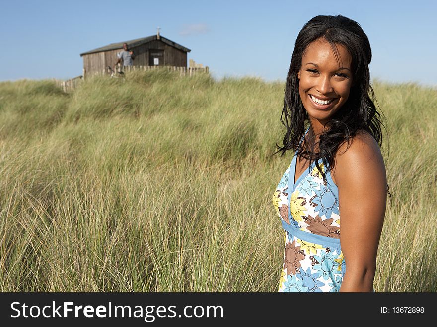 Young Woman Standing On Beach Amongst Dunes With Beach Hut In Distance