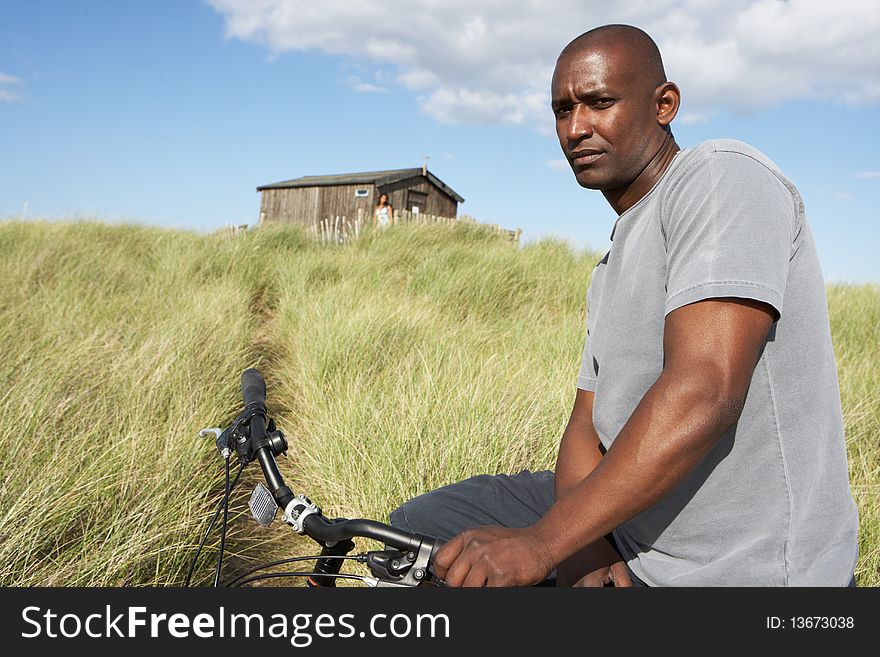 Young Man Riding Mountain Bike By Dunes With Old Beach Hut In Distance