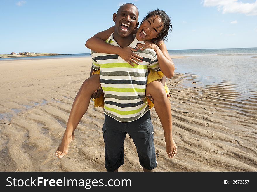 Young Man Giving Woman Piggyback Along Shoreline Of Beach