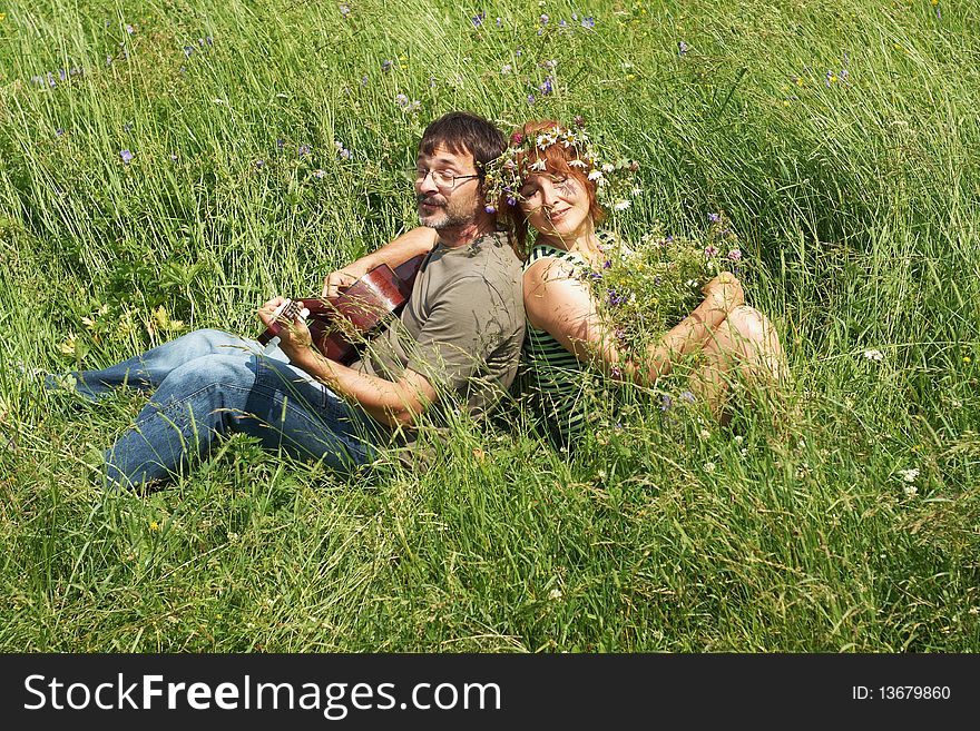 The pair sits on a meadow with a guitar. The pair sits on a meadow with a guitar