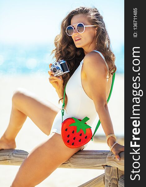 Trendy woman in white beachwear with retro photo camera sitting on a wooden fence on the seacoast. woman in UVA UVB protectant sunglasses with funny strawberry shape bag. Capture vacation. Trendy woman in white beachwear with retro photo camera sitting on a wooden fence on the seacoast. woman in UVA UVB protectant sunglasses with funny strawberry shape bag. Capture vacation