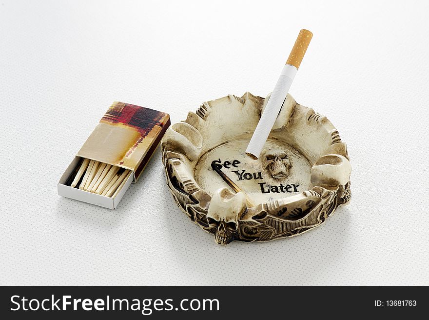 Cigarette in skull ashtray, with 'See You Later' motif and matches on a tabletop. Cigarette in skull ashtray, with 'See You Later' motif and matches on a tabletop