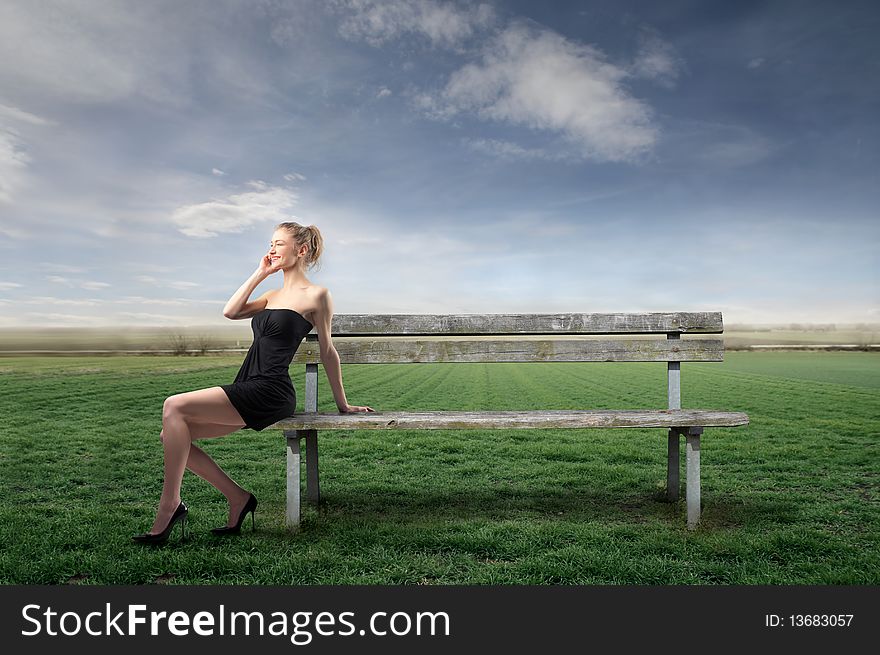 Elegant young woman using a mobile phone while sitting on a park bench on a green meadow. Elegant young woman using a mobile phone while sitting on a park bench on a green meadow