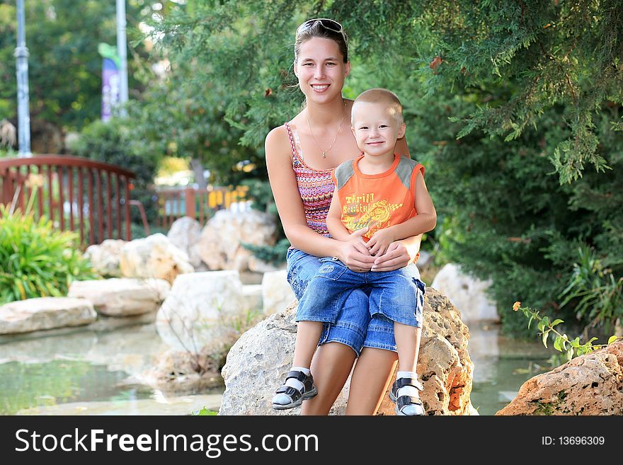 Young mother with her small child sitting on a large stone in a beautiful park and smiling. Young mother with her small child sitting on a large stone in a beautiful park and smiling