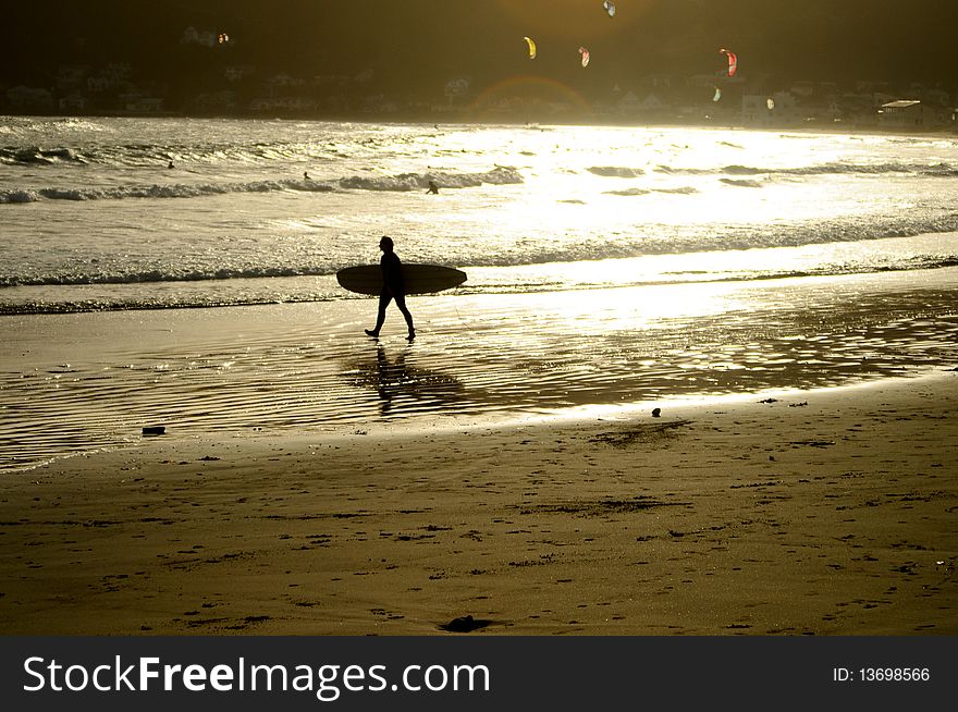 Surfer walking out to the waves at dusk with kite surfers in the background