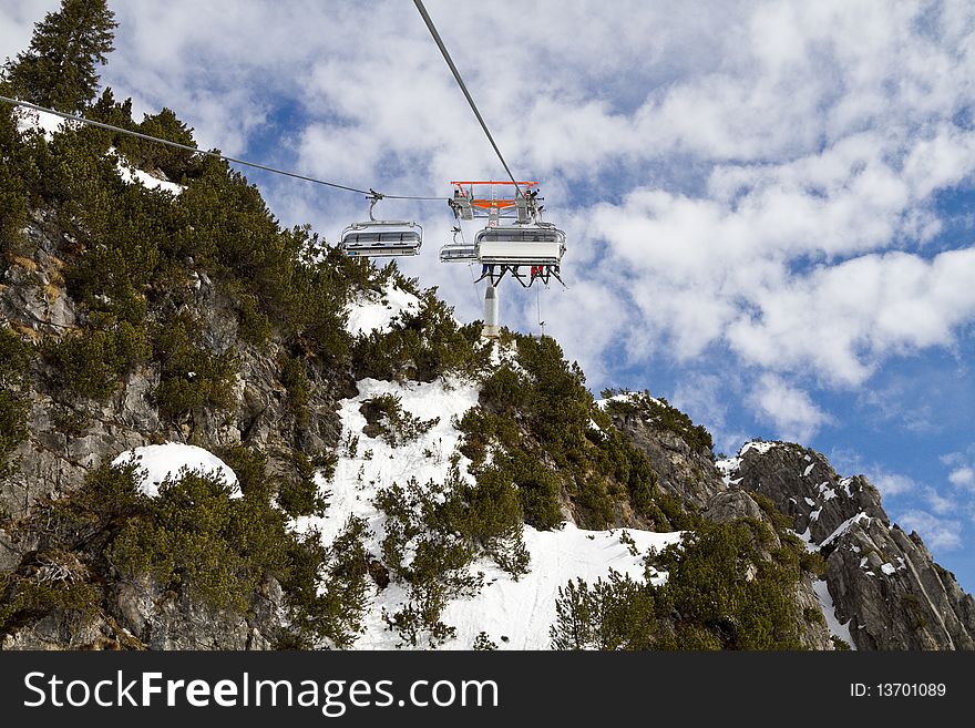 Ski chair lift with people above snowy rocky mountain against a cloudy blue sky. Ski chair lift with people above snowy rocky mountain against a cloudy blue sky