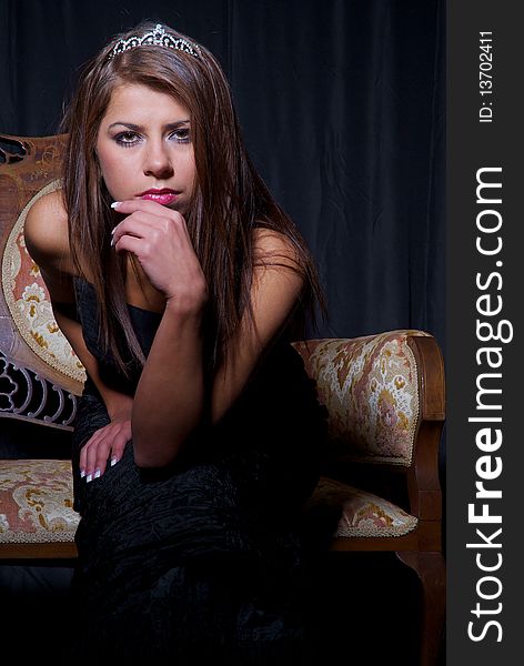 Girl wearing black prom dress and a crown sitting on the antique sofa, black background. Girl wearing black prom dress and a crown sitting on the antique sofa, black background