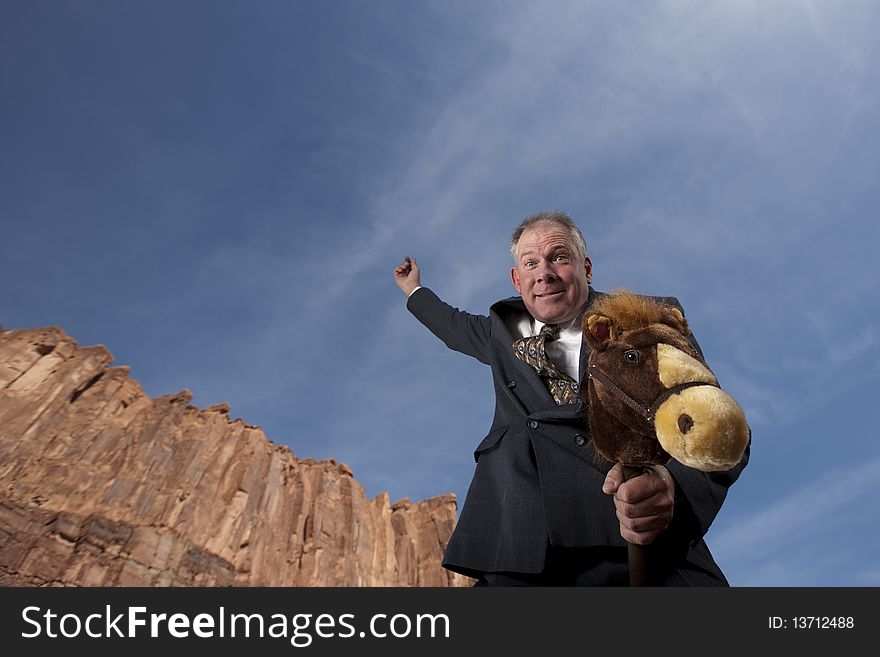 A smiling businessman rides a stick horse with one arm pointing to the sky in a desert landscape. Horizontal shot. A smiling businessman rides a stick horse with one arm pointing to the sky in a desert landscape. Horizontal shot.